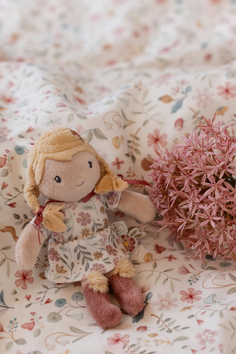 Boneca de peluche com vestido floral sentada sobre tecido branco floral ao lado de flores cor-de-rosa