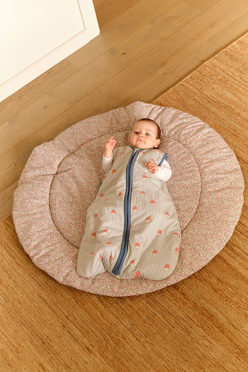 Bebé deitado numa almofada redonda floral com saco de dormir cinzento às riscas.