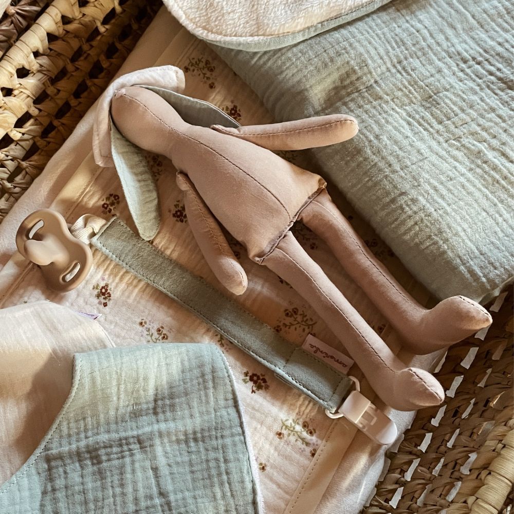 Boneca de tecido bege com orelhas verdes em berço com colcha estampada, almofada verde, chupeta bege e mordedor com fita e etiqueta.