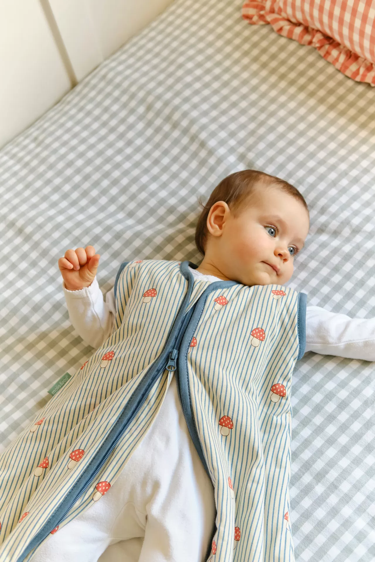 Bébé deitado num colchão xadrez cinzento e branco com saco de dormir azul claro com cogumelos vermelhos