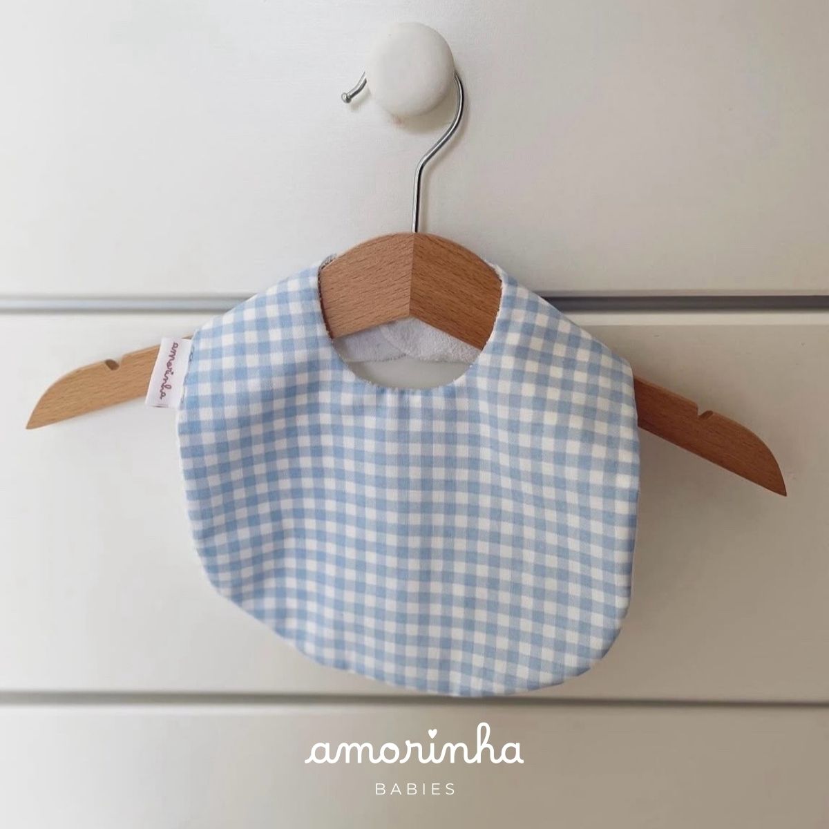 Babete de bebé azul e branco com padrão quadriculado pendurado num cabide de madeira.