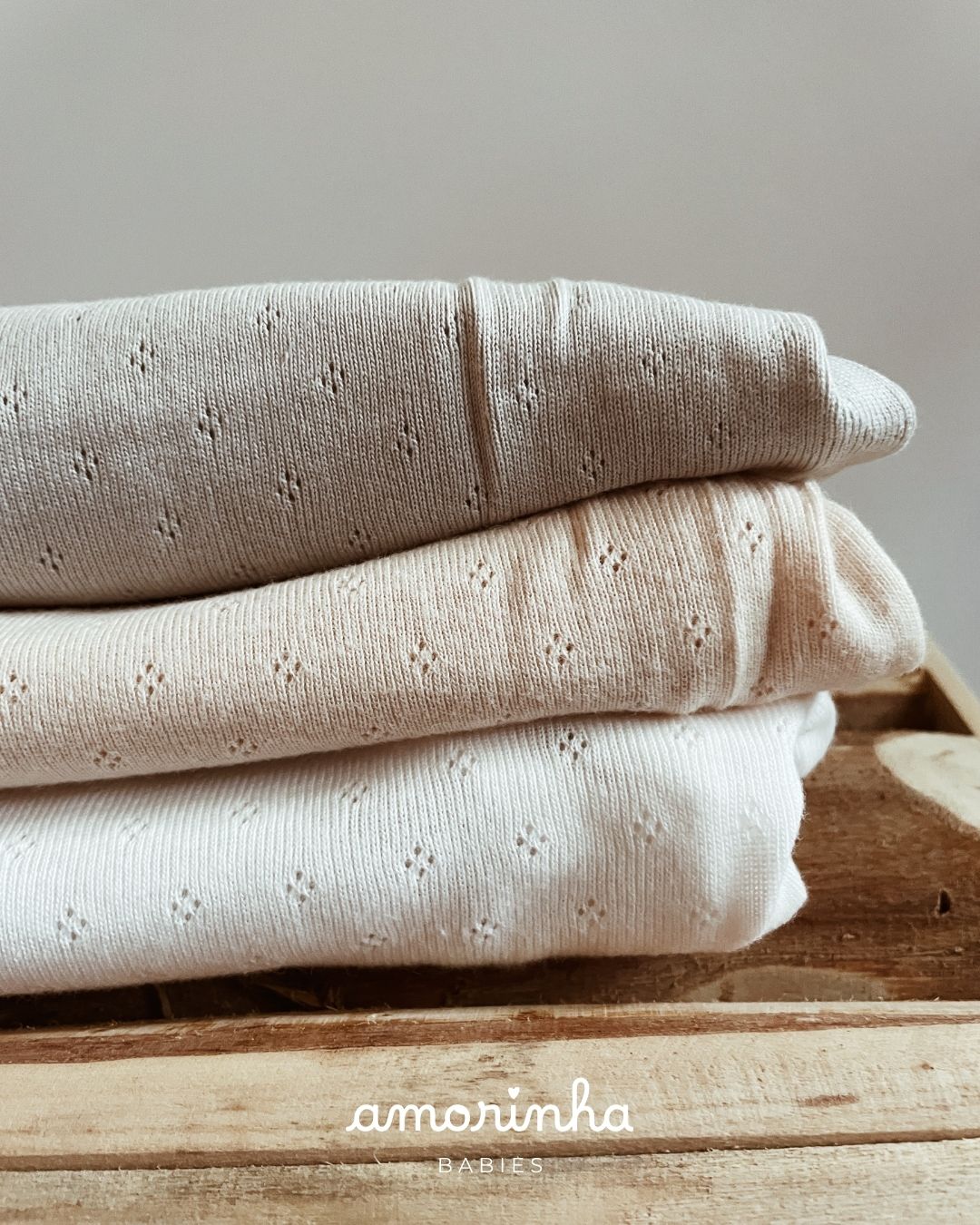 Peças de roupa em tons neutros com padrão de pequenos furos sobre caixa de madeira com texto