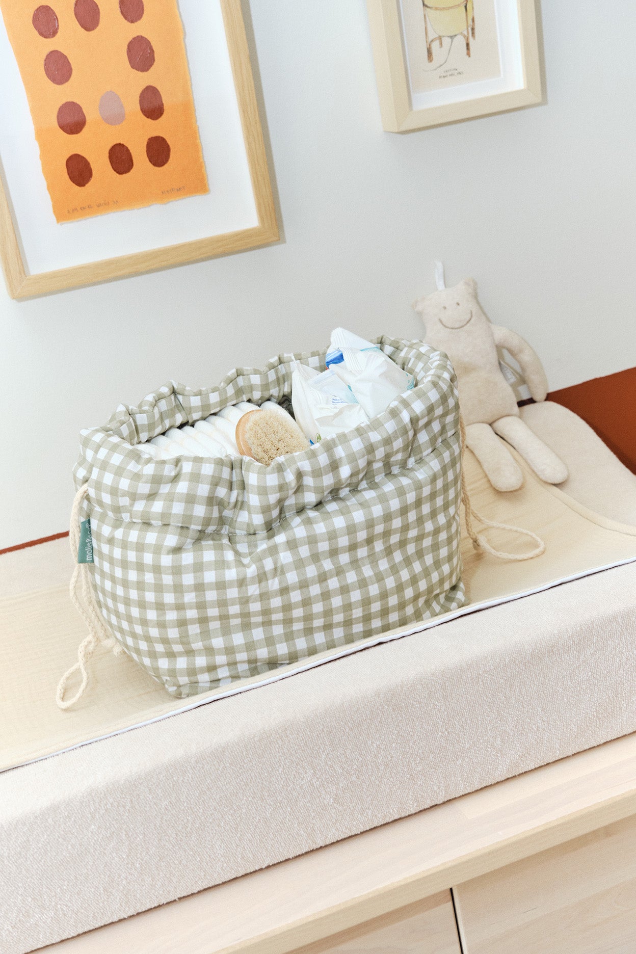 Bolsa organizadora xadrez verde claro com fraldas, lenços e escova, sobre trocador com peluche ao fundo.