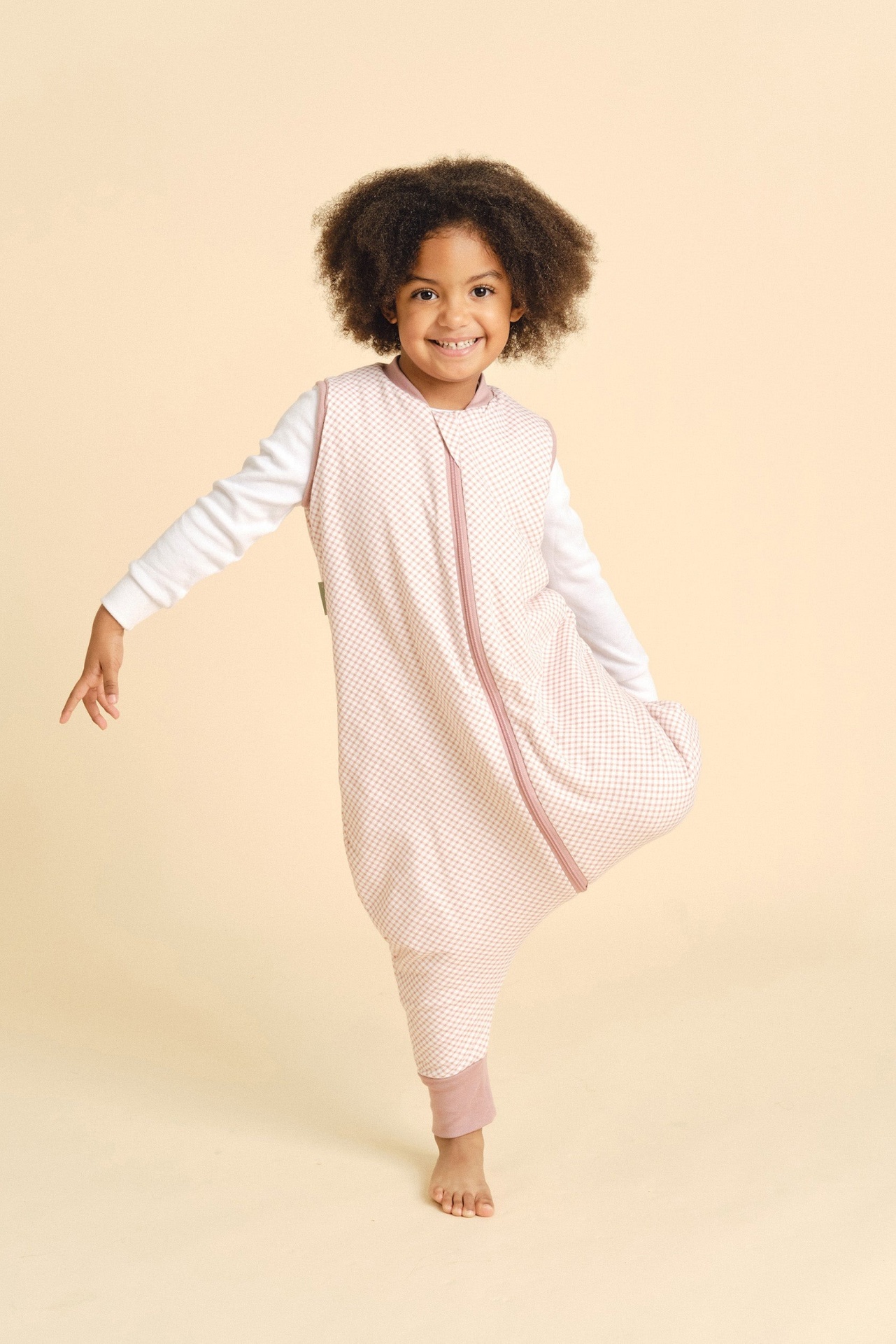 Menina com pijama de padrão xadrez rosa e branco e camisa branca, com sorriso, fundo bege claro