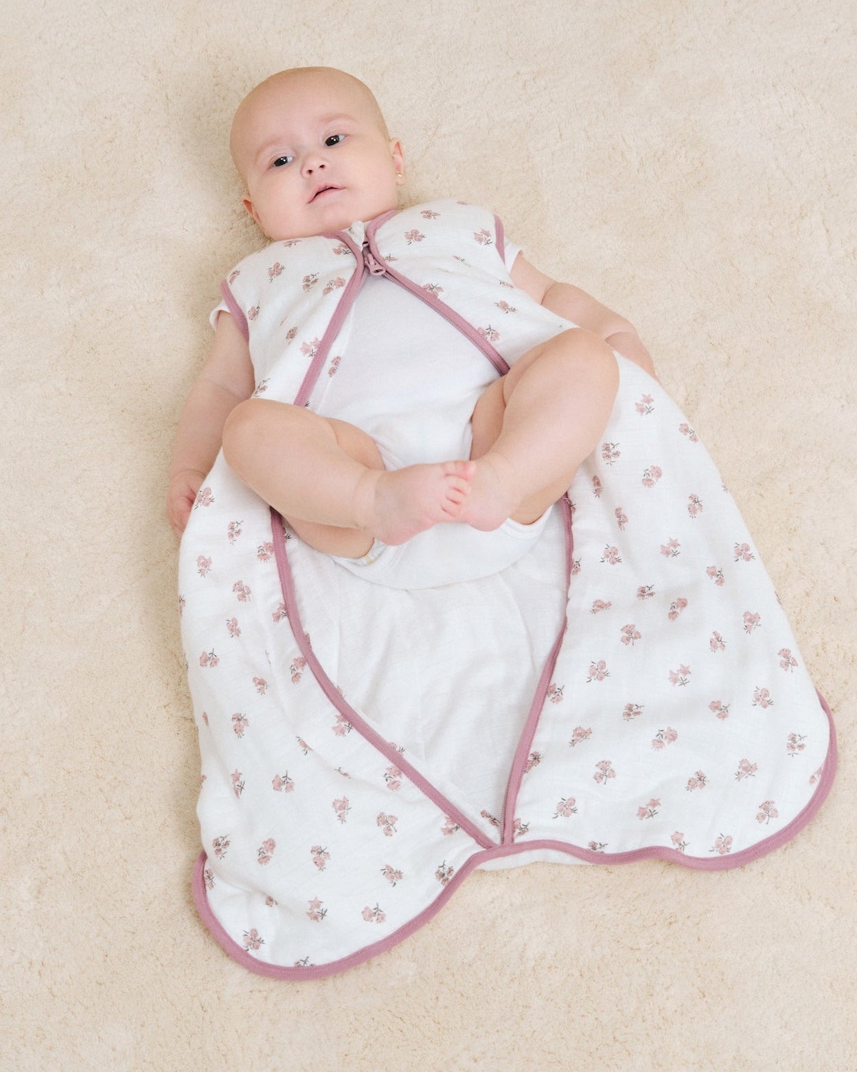 Bebé deitado com saco de dormir branco com flores cor-de-rosa e fecho de correr cor-de-rosa