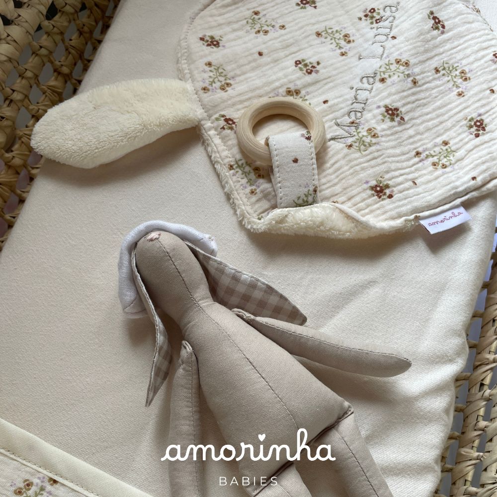 Brinquedo coelho de peluche bege e brinquedo mordedor floral com argola de madeira em berço de palha.