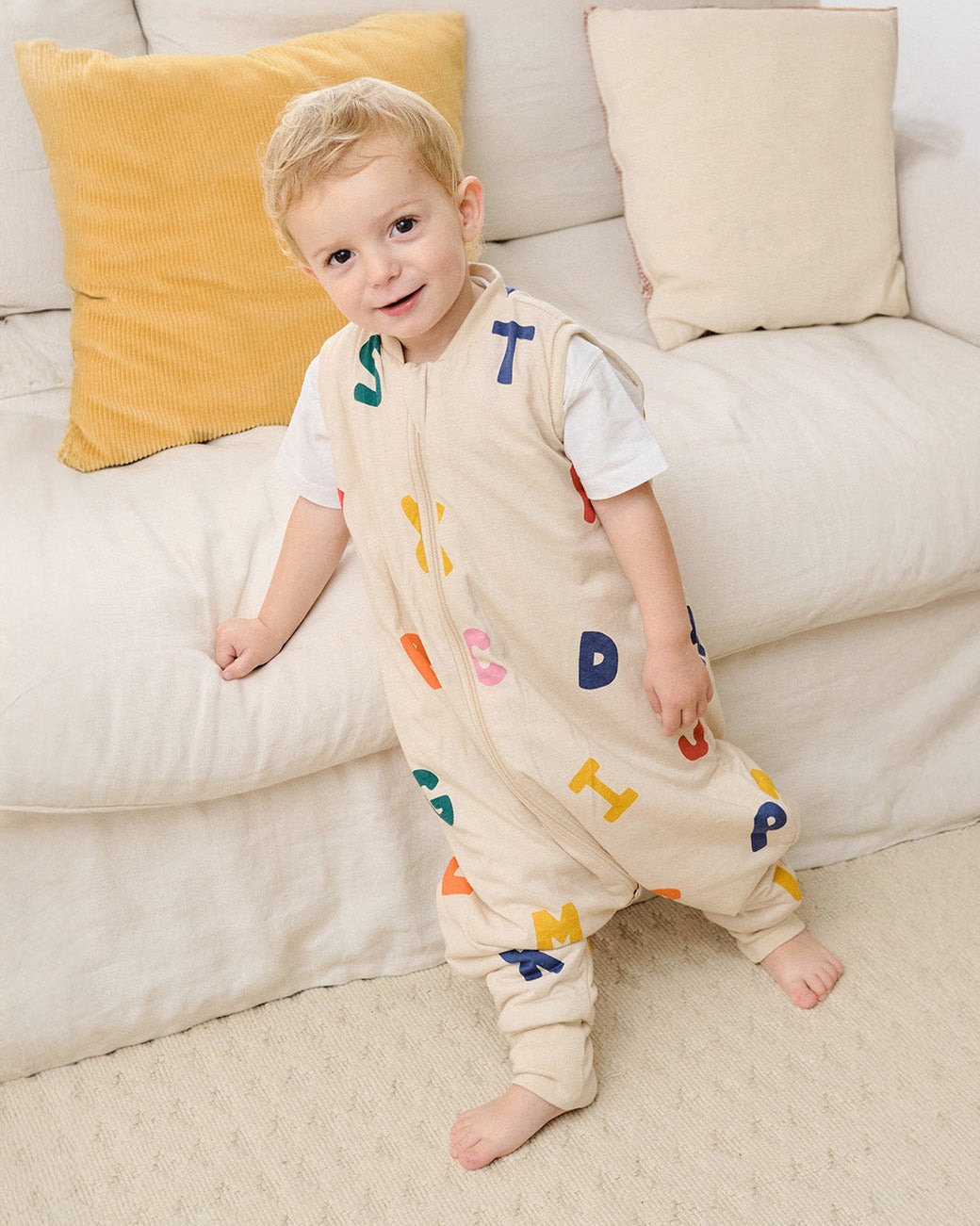 Criança com pijama saco de dormir bege com letras coloridas sentado num sofá creme com almofadas
