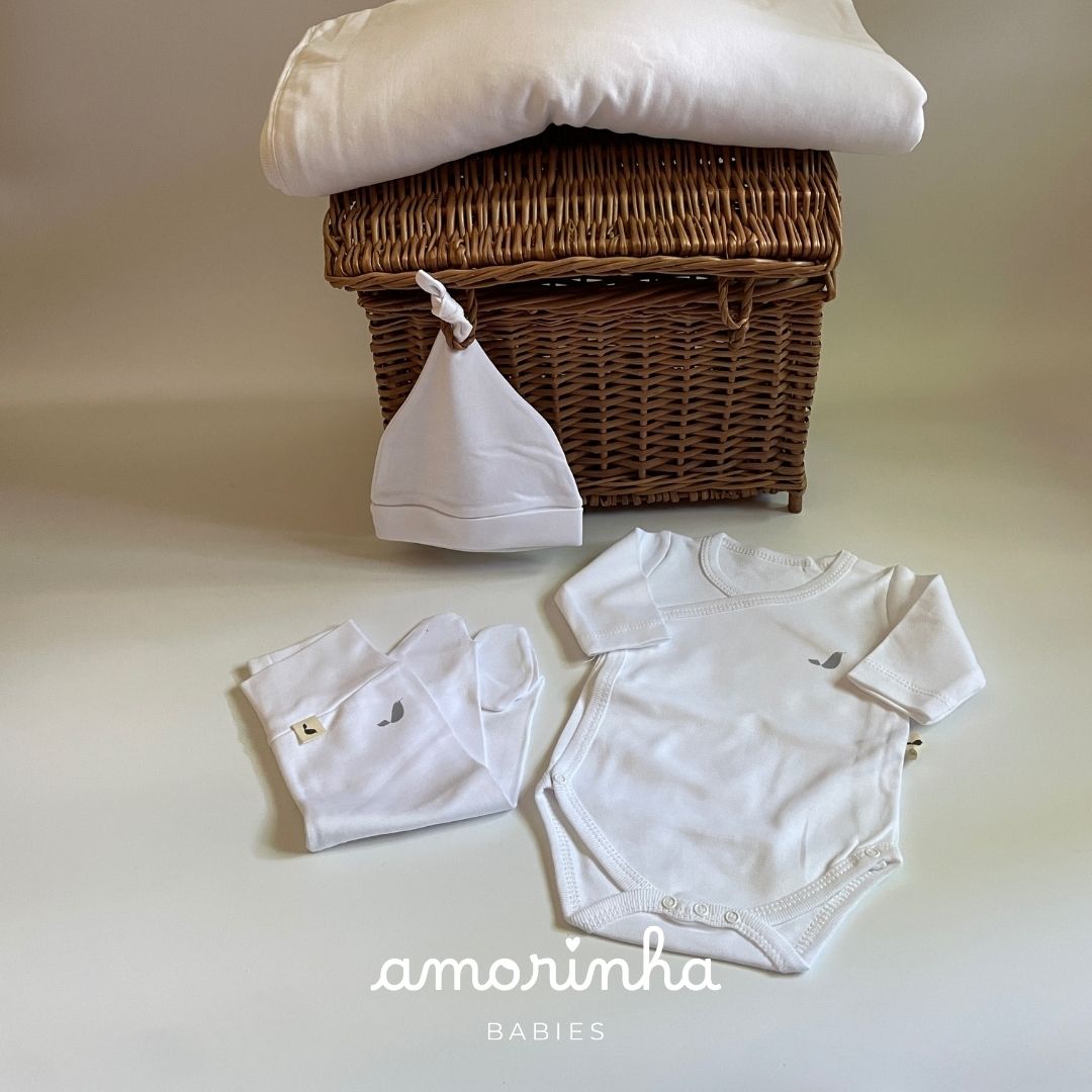 Conjunto de roupa de bebé branca com body, calças e gorro em frente a um cesto de vime e cobertor branco
