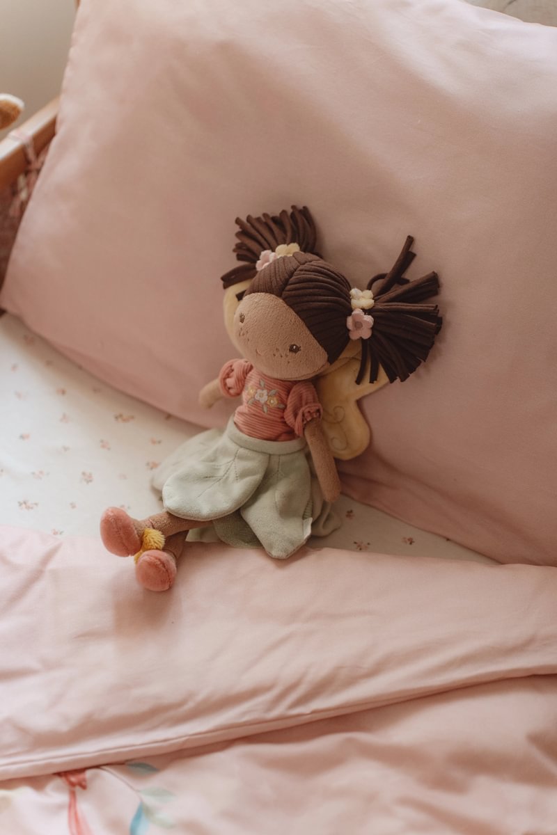 Boneca de pano com roupa cor-de-rosa e saia clara numa cama com roupa de cama rosa