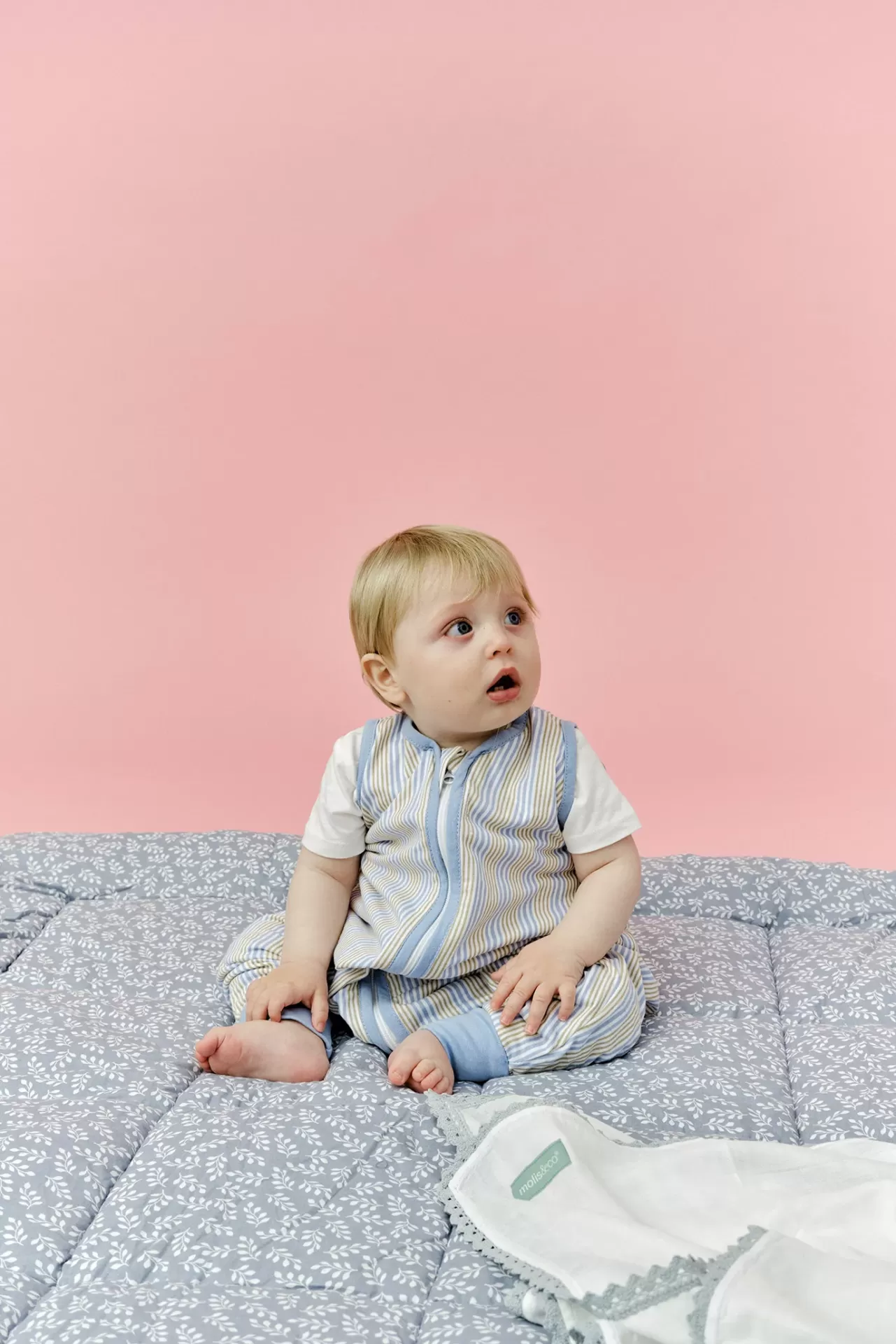 Bebé vestido com roupa às riscas sentado num colchão cinza com padrão de folhas e manta branca com renda