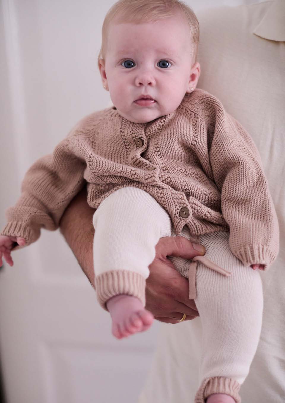 Bebé com casaco e calças de malha em tons bege e branco a ser segurado por adulto