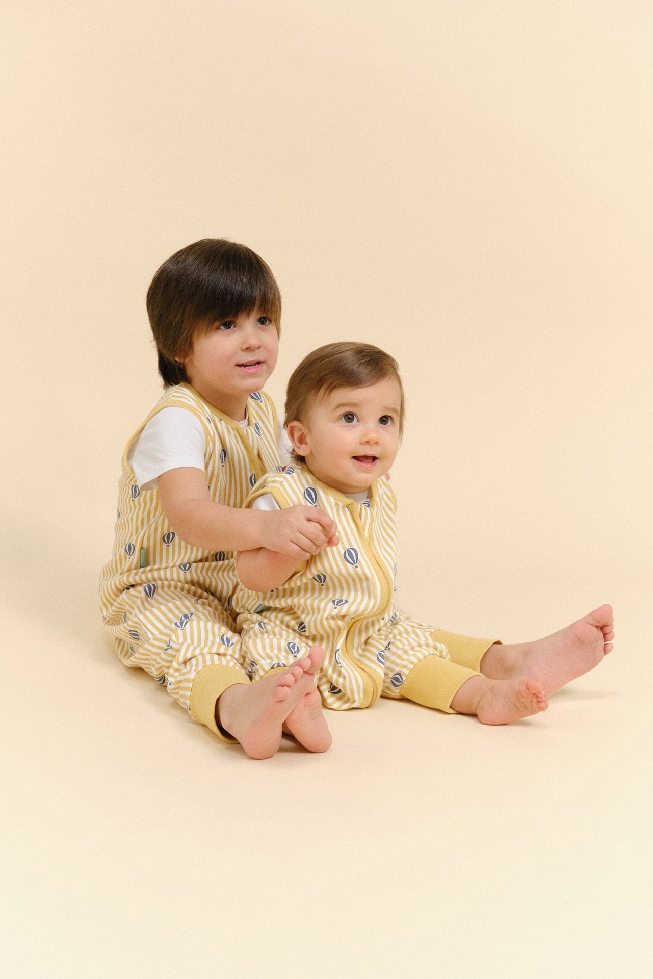 Dois bebés com macacões amarelos às riscas e balões azuis sentados no chão creme