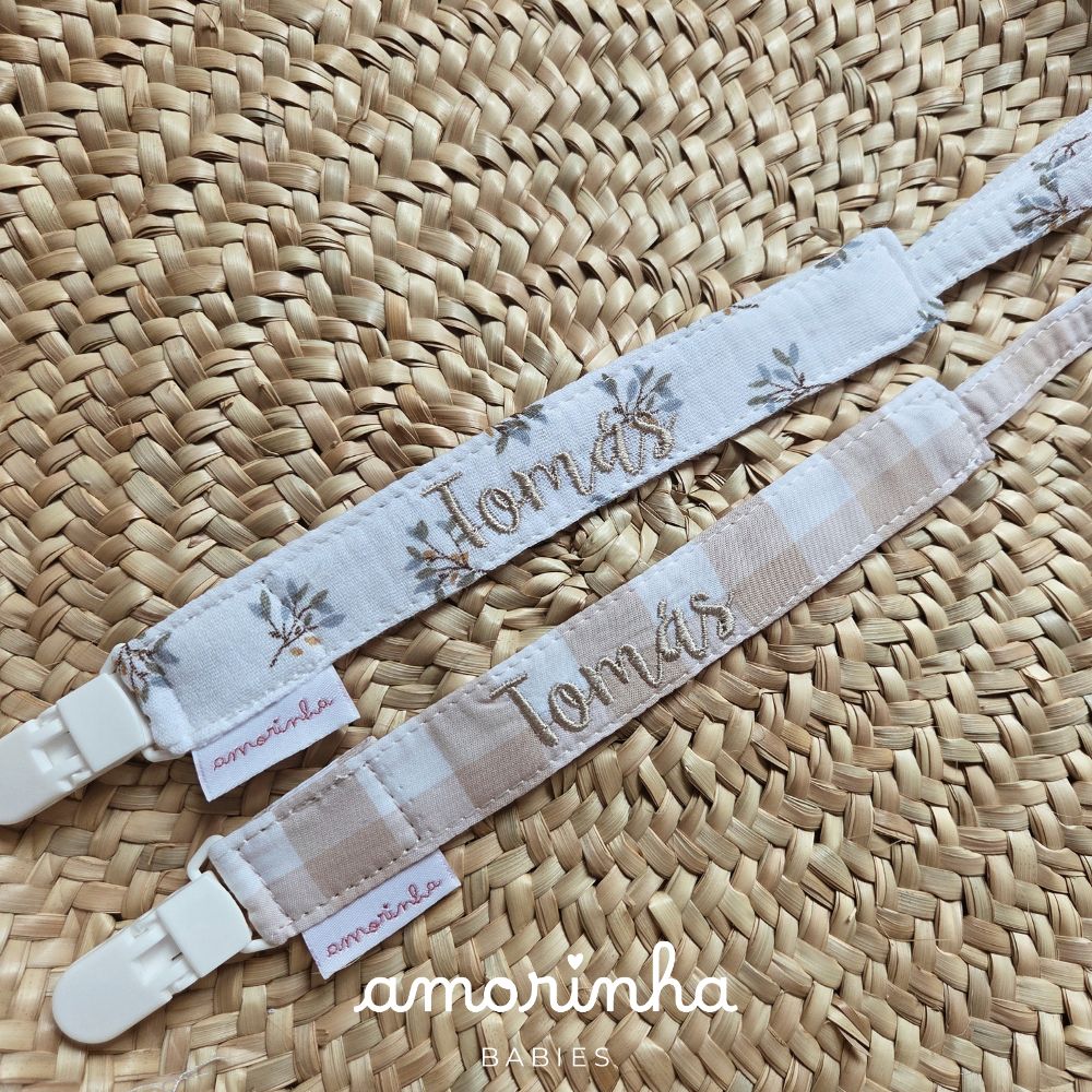Duas fitas para chupeta com nome Tomás bordado, padrões floral e xadrez, sobre tampa em palha entrelaçada.