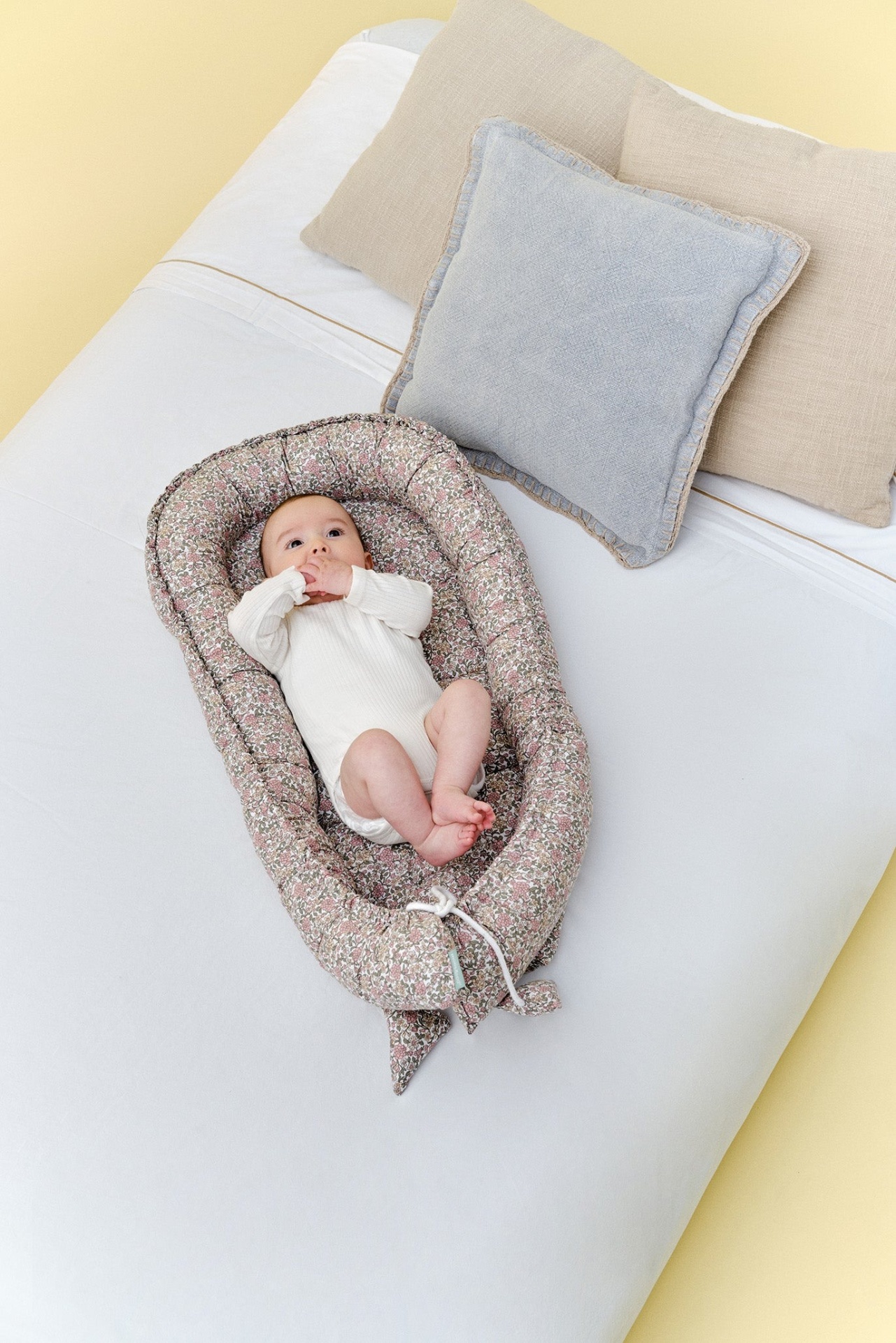 Berço portátil floral acolchoado com bebé deitado sobre colchão branco e almofadas.
