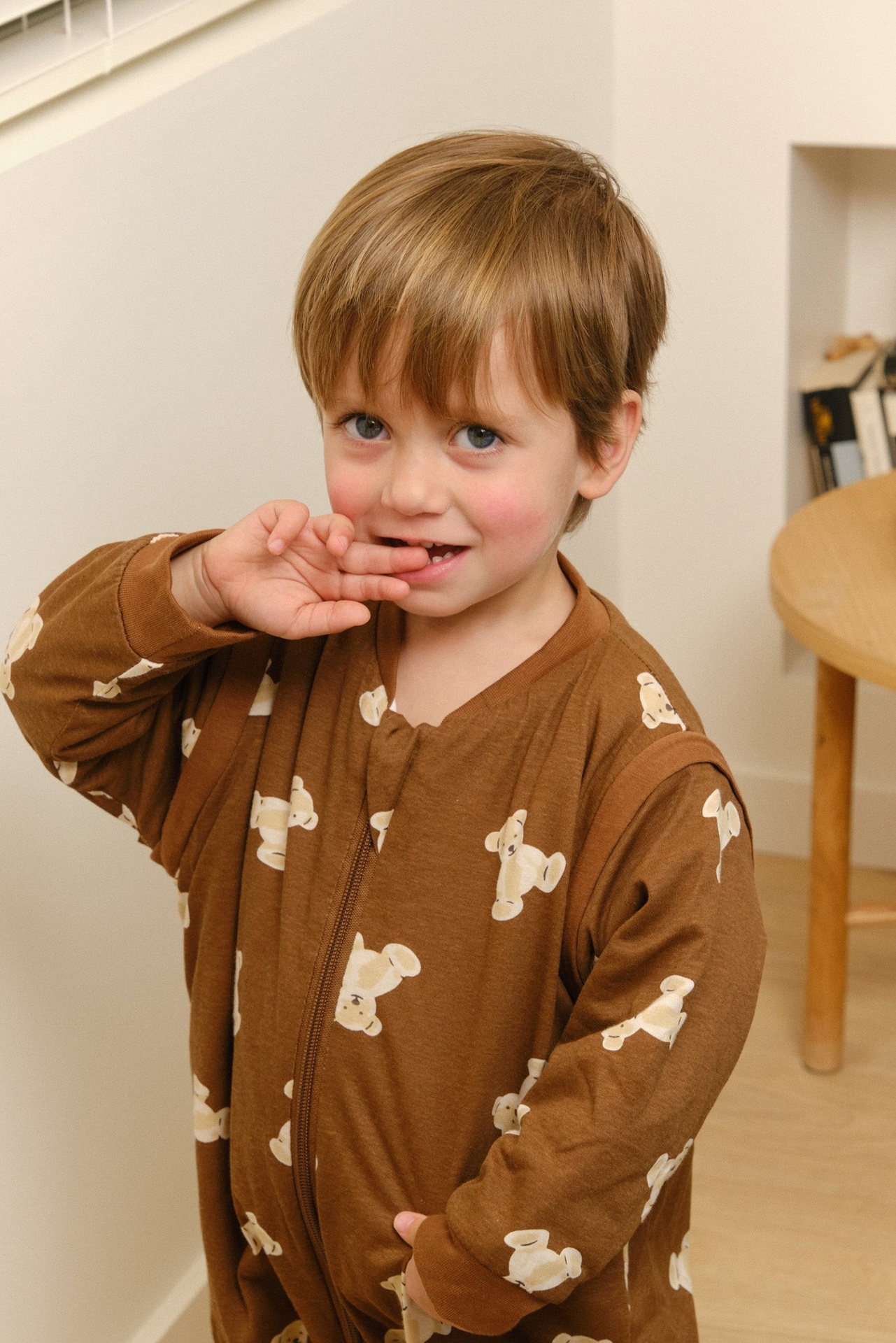 Criança em pijama castanho com ursinhos creme em ambiente interior com parede branca e mesa em madeira