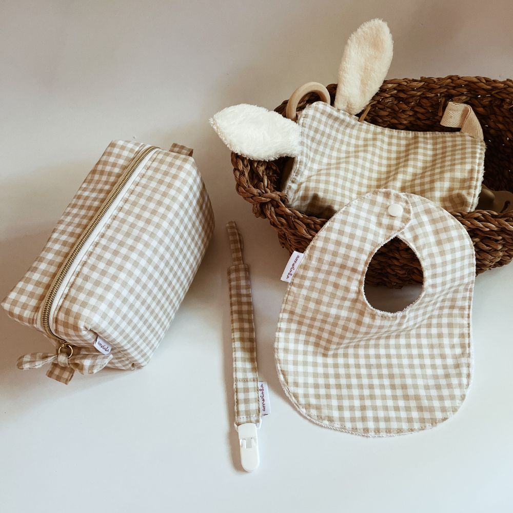 Acessórios de bebé em tecido xadrez bege e branco sobre mesa branca com cesta de vime