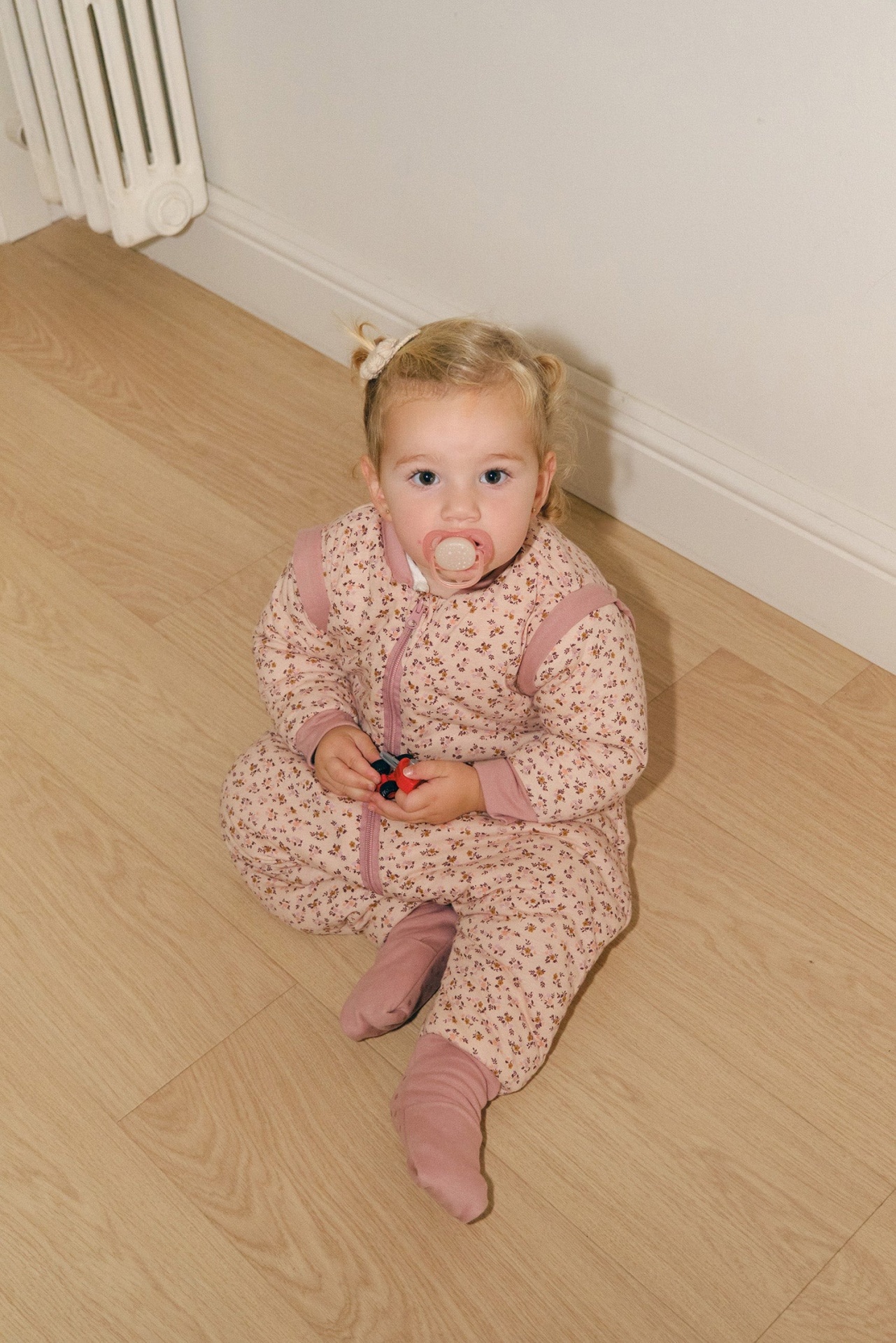 Bebé com pijama rosa floral, chupeta e brinquedo sentado no chão de madeira clara
