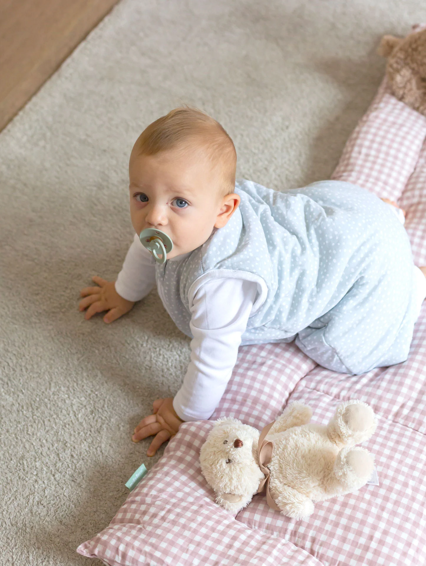 Bebé com saco de dormir azul de bolinhas em manta rosa quadriculada com ursinho de peluche