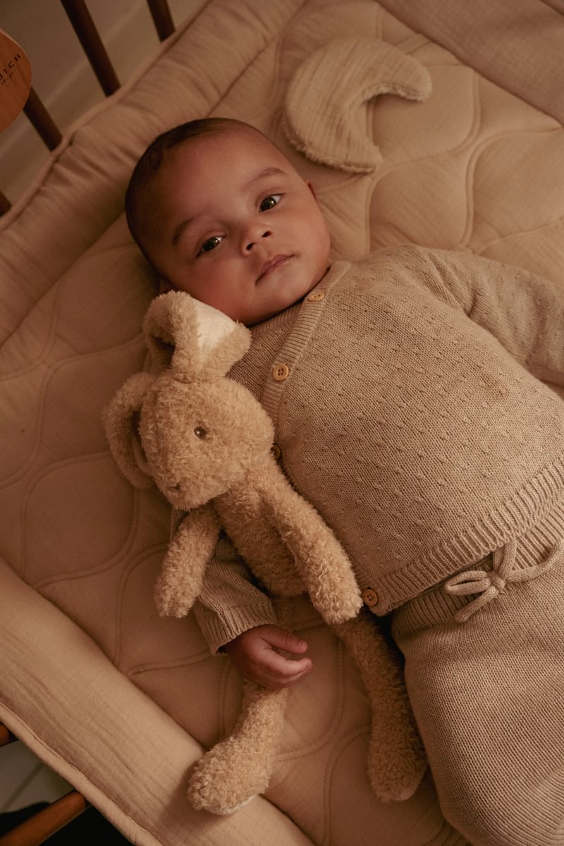 Bebé vestido com conjunto de malha bege deitado em cama com edredão bege segurando peluche castanho de coelho.