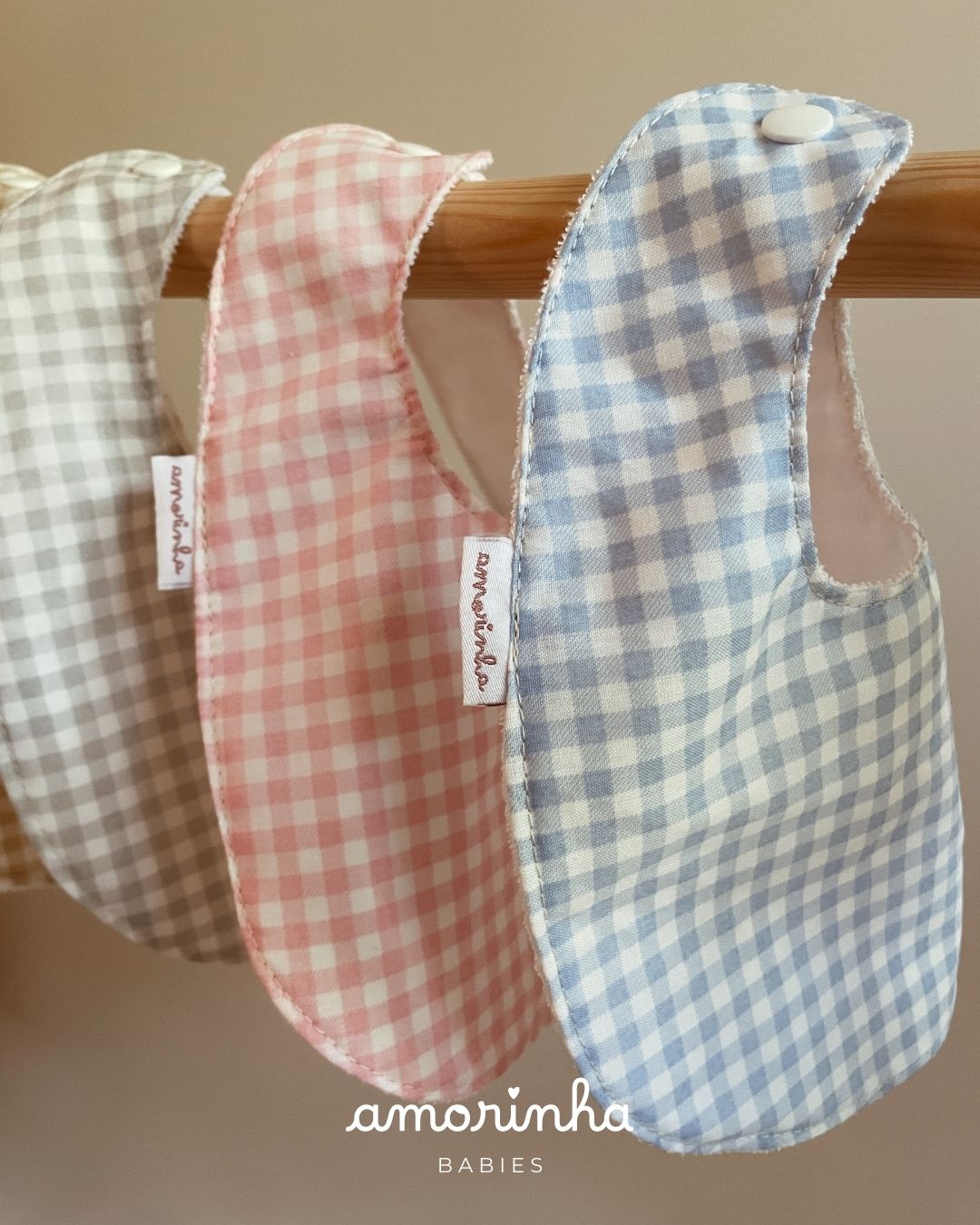 Três babetes xadrez para bebés em azul, rosa e cinza pendurados numa vara de madeira