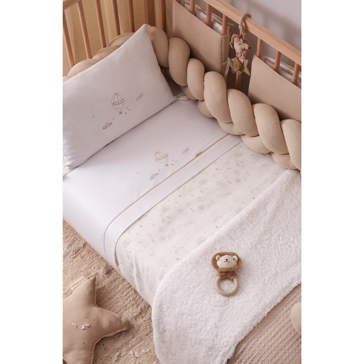 Berço de bebé com roupa de cama branca e bege, protetor trançado, peluche de leão e almofada estrela.