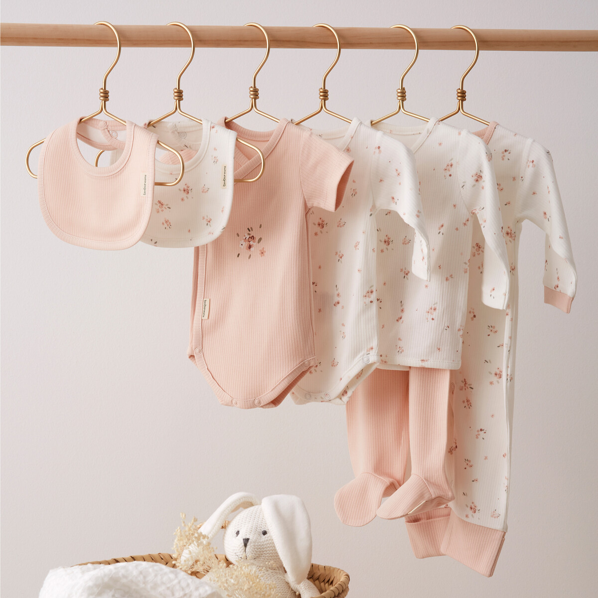 Roupas de bebé em rosa e branco com padrão floral penduradas numa barra de madeira.