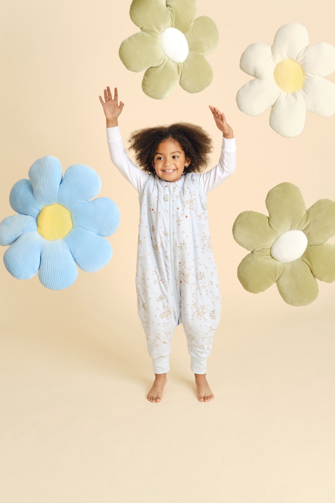 Criança com macacão floral branco com almofadas em forma de flor azul, verde e branca em fundo bege.