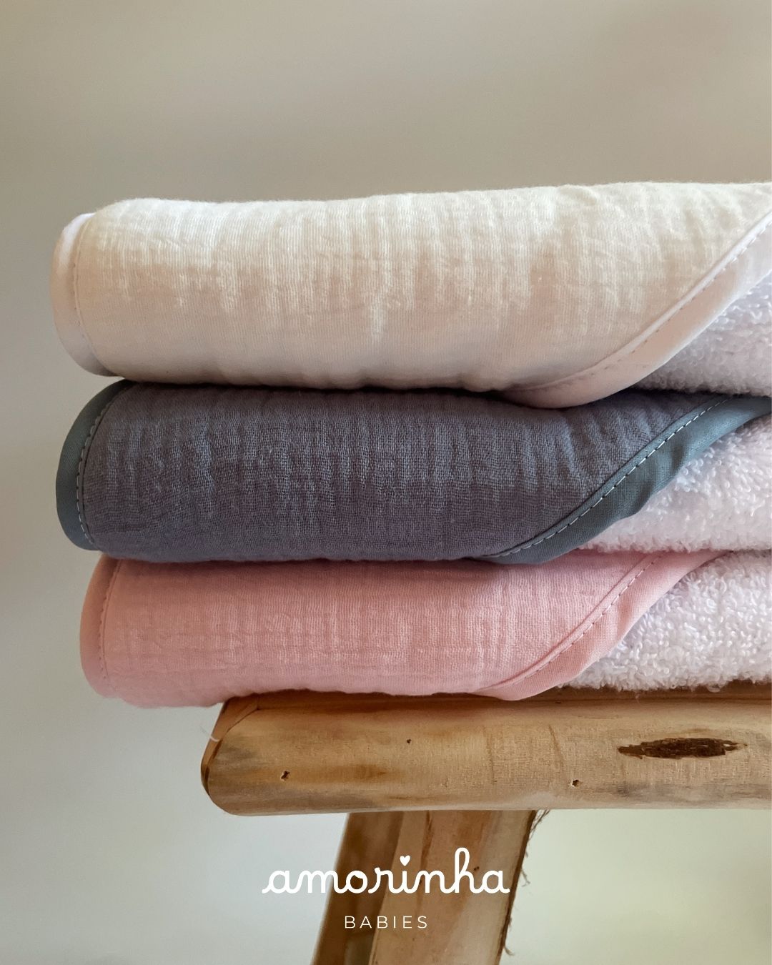 Toalhas de bebé em branco, cinza e rosa empilhadas sobre uma cadeira de madeira.