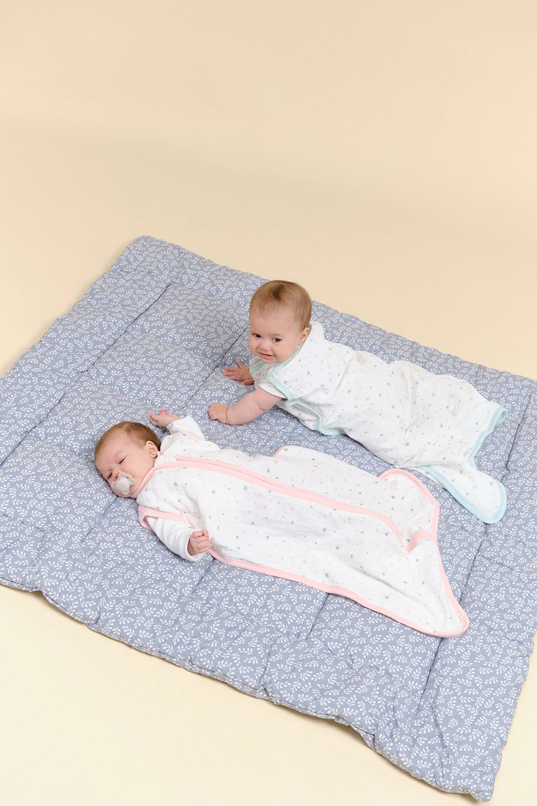 Dois bebés em saquinhos de dormir brancos com estrelas sobre tapete azul acolchoado com padrão de folhagem.