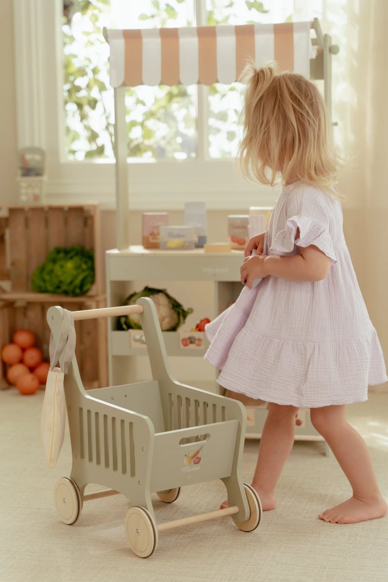 Carrinho de madeira cinza claro para crianças ao lado de menina com vestido branco lilás e loja de brinquedo
