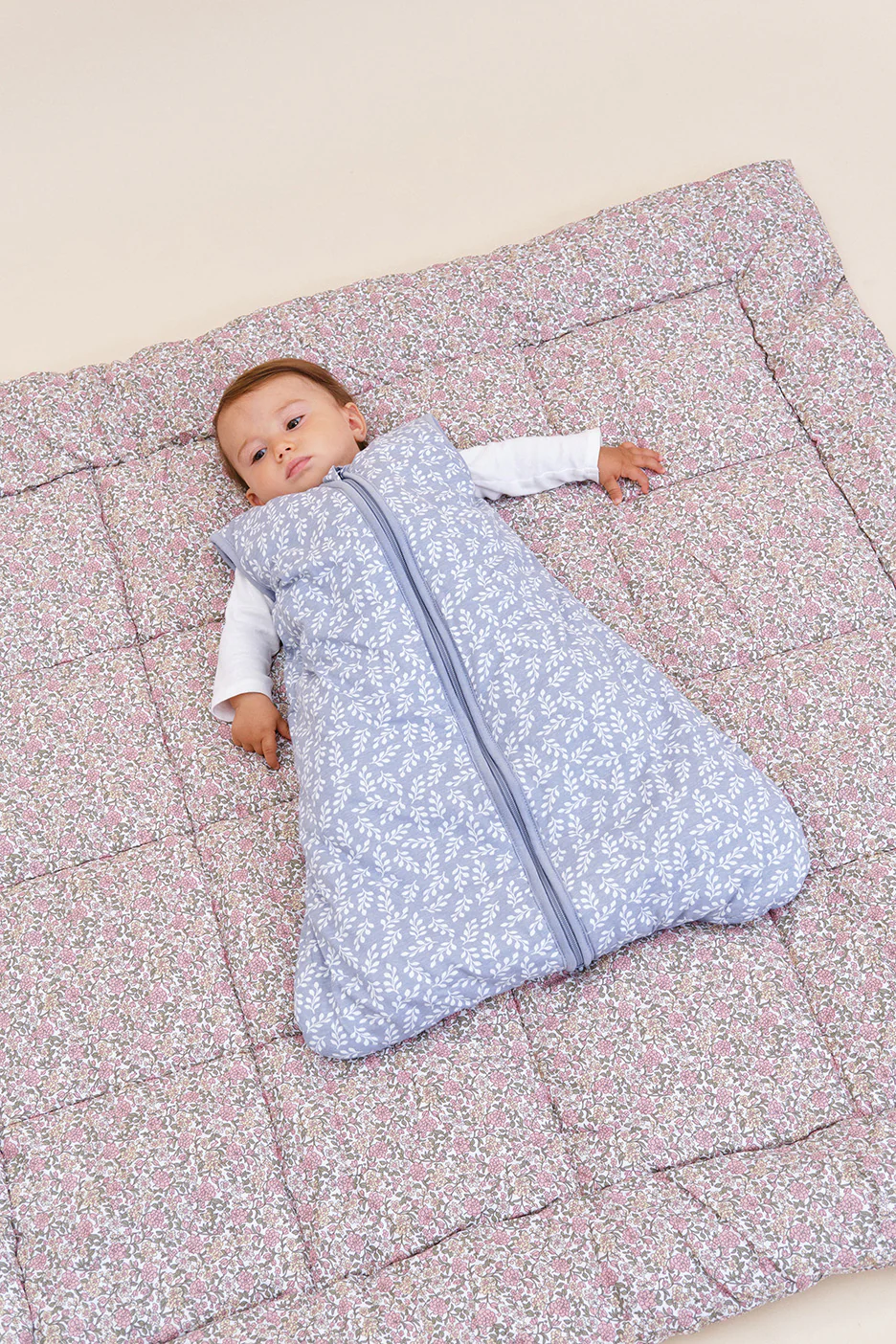 Bebé deitado em saco de dormir azul com padrão branco sobre edredom floral rosa.