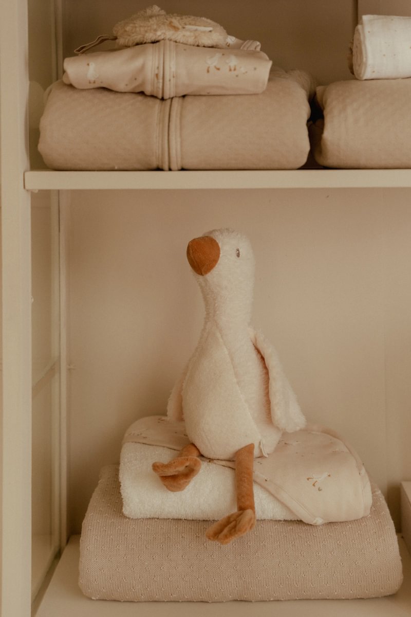 Peluche de pato macio em prateleira com mantas e roupões beges e brancos.