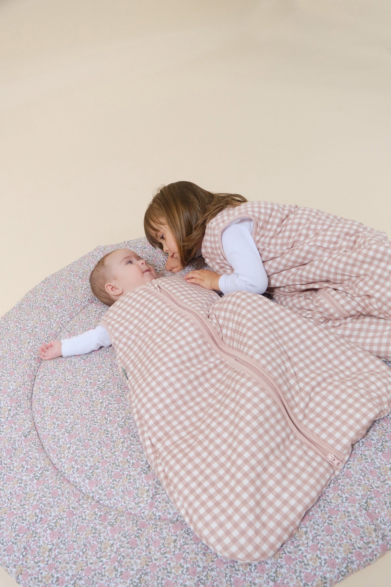 Dois bebés com sacos de dormir xadrez rosa e branco num tapete floral redondo