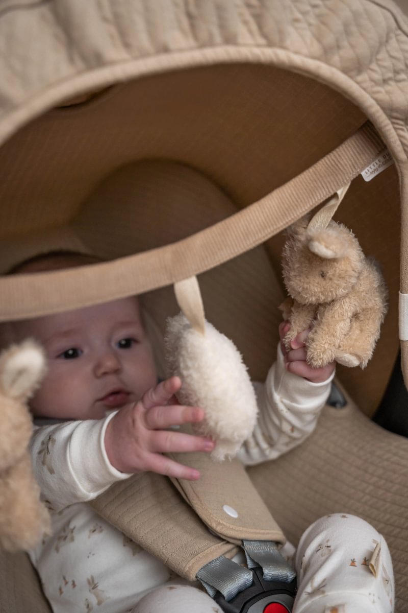 Bebé com roupa clara a brincar com brinquedos de peluche numa cadeirinha de bebé bege acolchoada