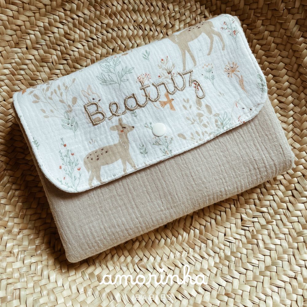 Carteira de tecido com aba estampada e nome bordado sobre fundo de palha trançada.