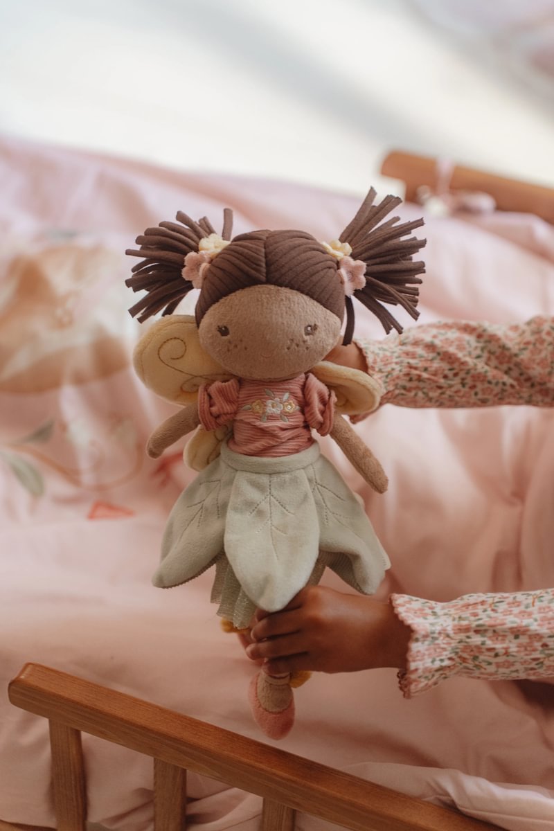 Boneca de pano fada com cabelo castanho e vestido rosa segurada por criança numa cama rosa