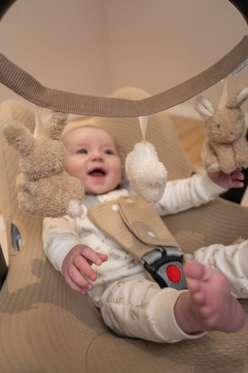 Bebé no cadeirão bege com mó de brinquedos pendurados castanhos e brancos