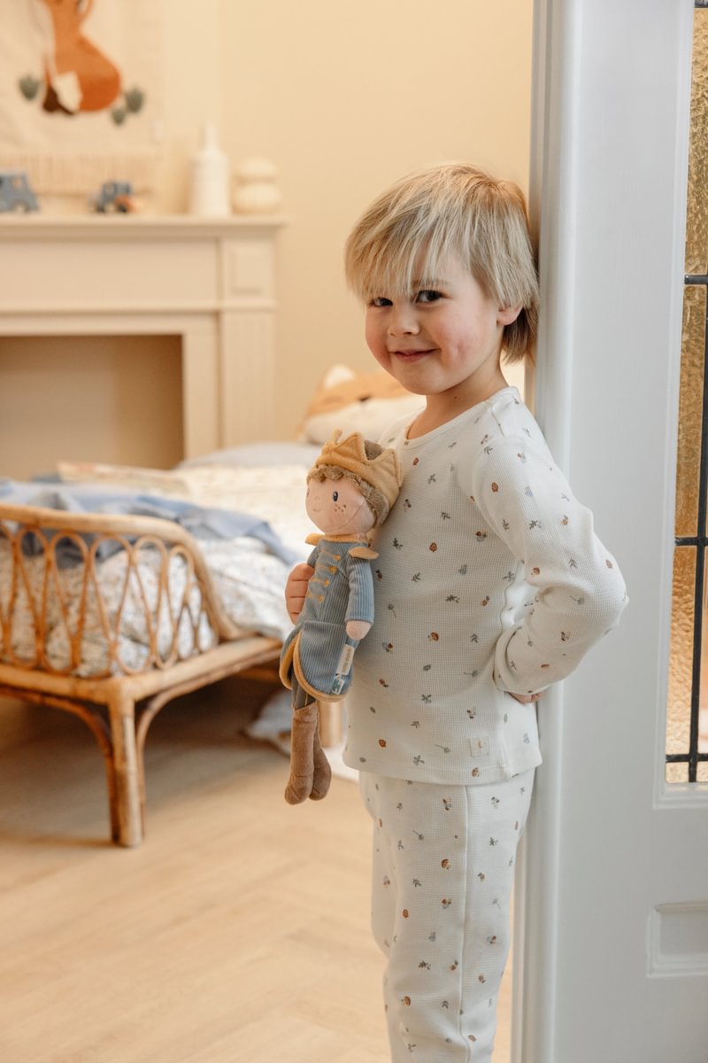 Menino loiro com pijama branco com padrão colorido segurando boneca de pano azul junto a porta