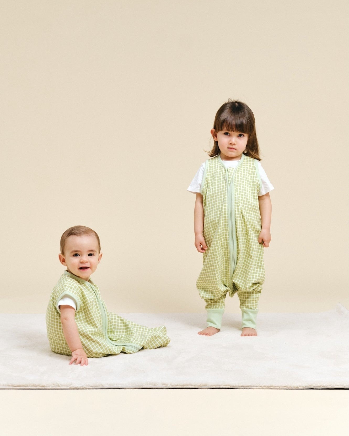 Duas crianças em pijamas saco verde claro com padrão de quadrados em fundo bege.