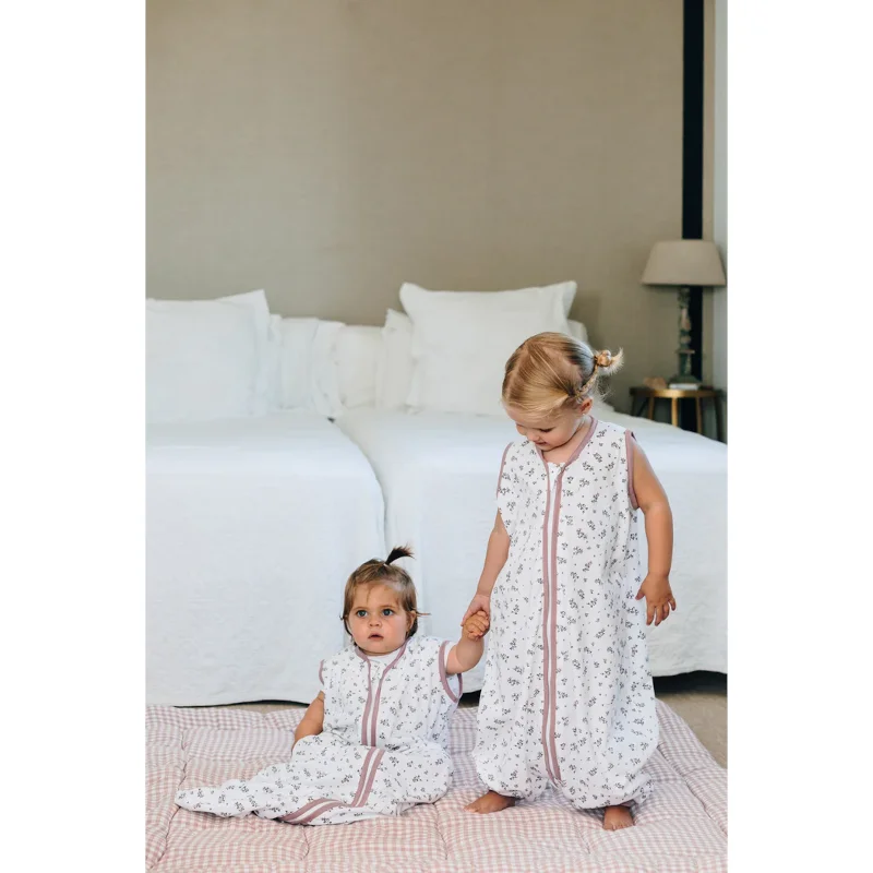 Dois bebés com sacos de dormir brancos com padrão floral e fecho rosa, numa manta rosa claro, num quarto com cama branca.