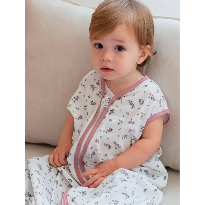Bebé com saco de dormir branco com padrão de flores e fecho rosa sentado num sofá bege