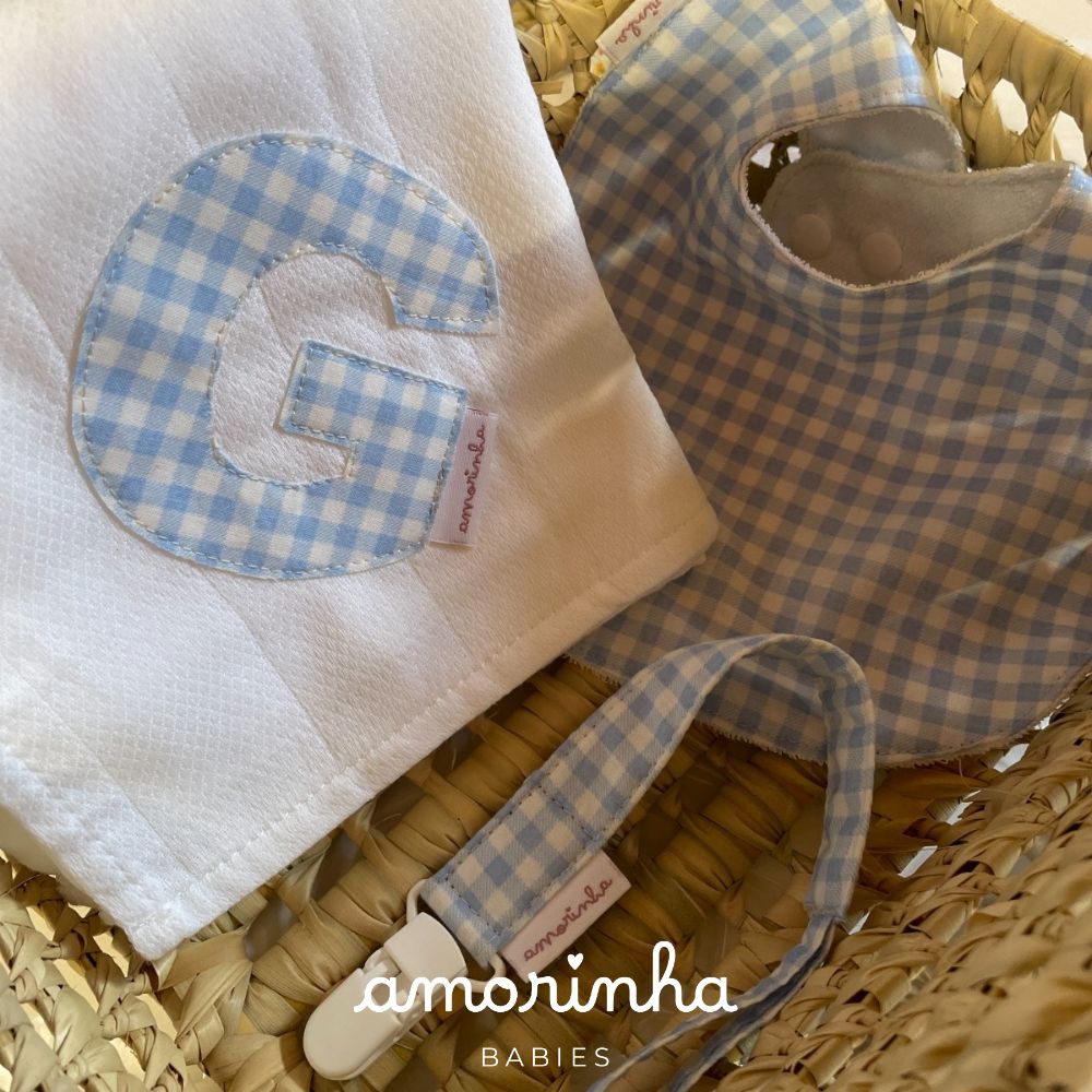 Conjunto de bebé branco e azul com babete, fralda e clip, etiquetas amorinha, em cesta de palha