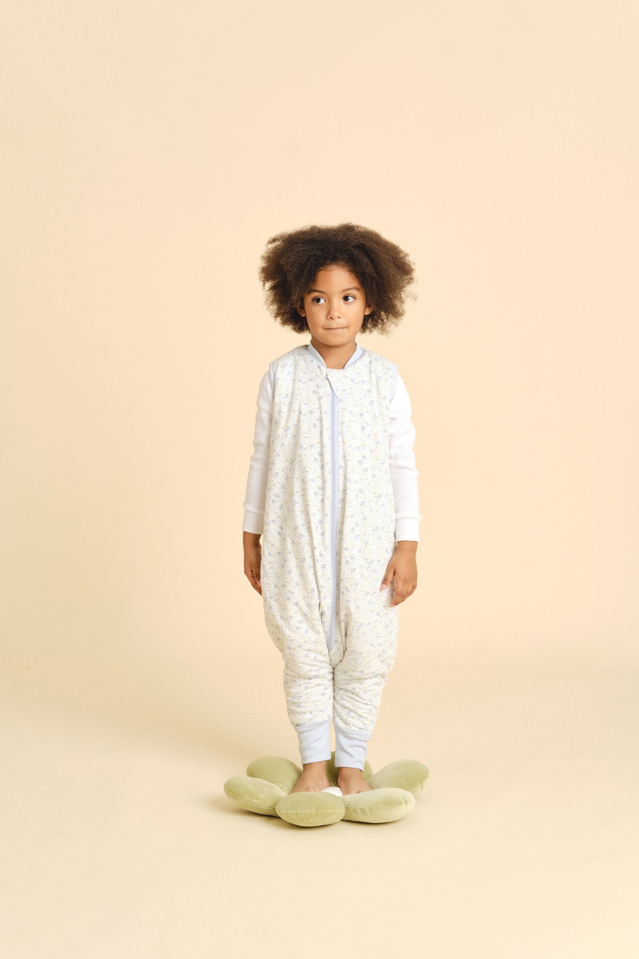Menina com saco de dormir acolchoado com padrão floral e camisola branca num fundo bege a pisar almofada verde em forma de flor.