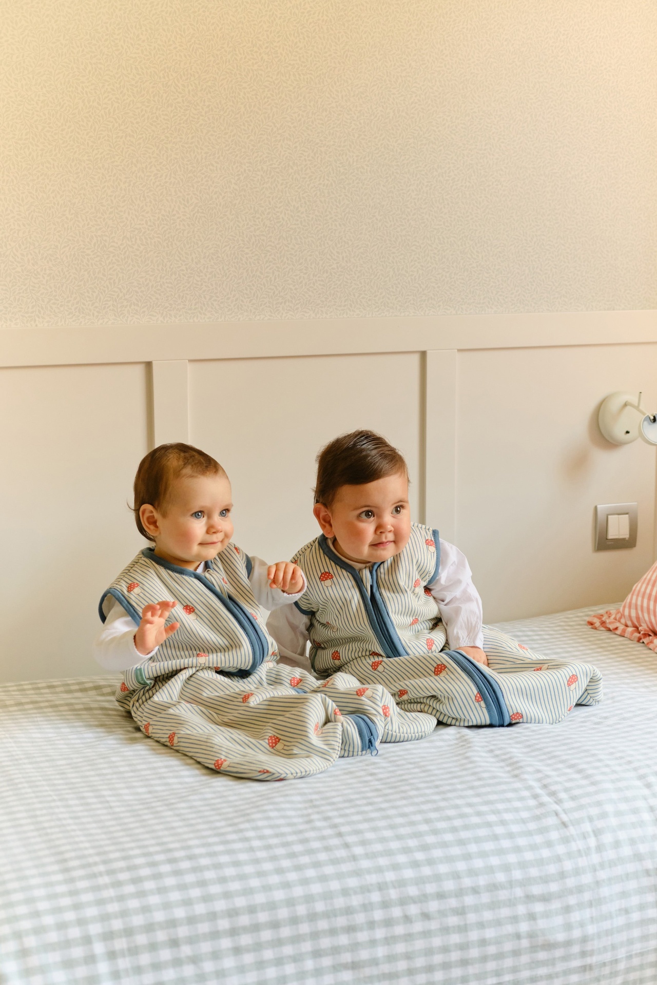 Dois bebés com sacos de dormir às riscas sentados numa cama com lençol xadrez verde claro e branco.