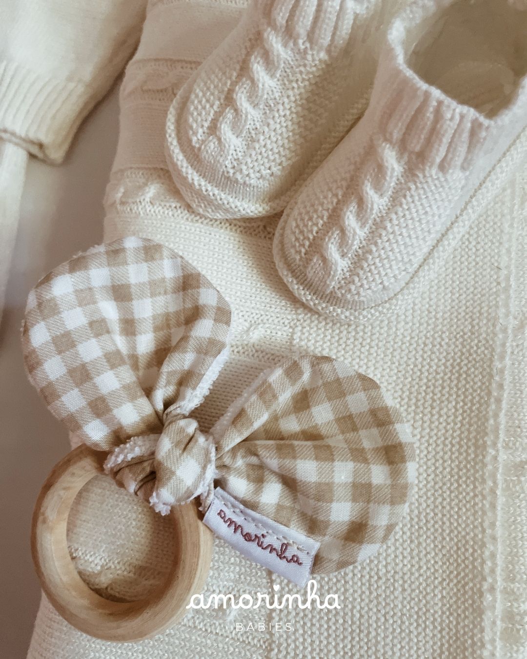 Chupeta de bebé de tecido xadrez bege e branco com argola de madeira sobre roupa de malha branca com pantufas.