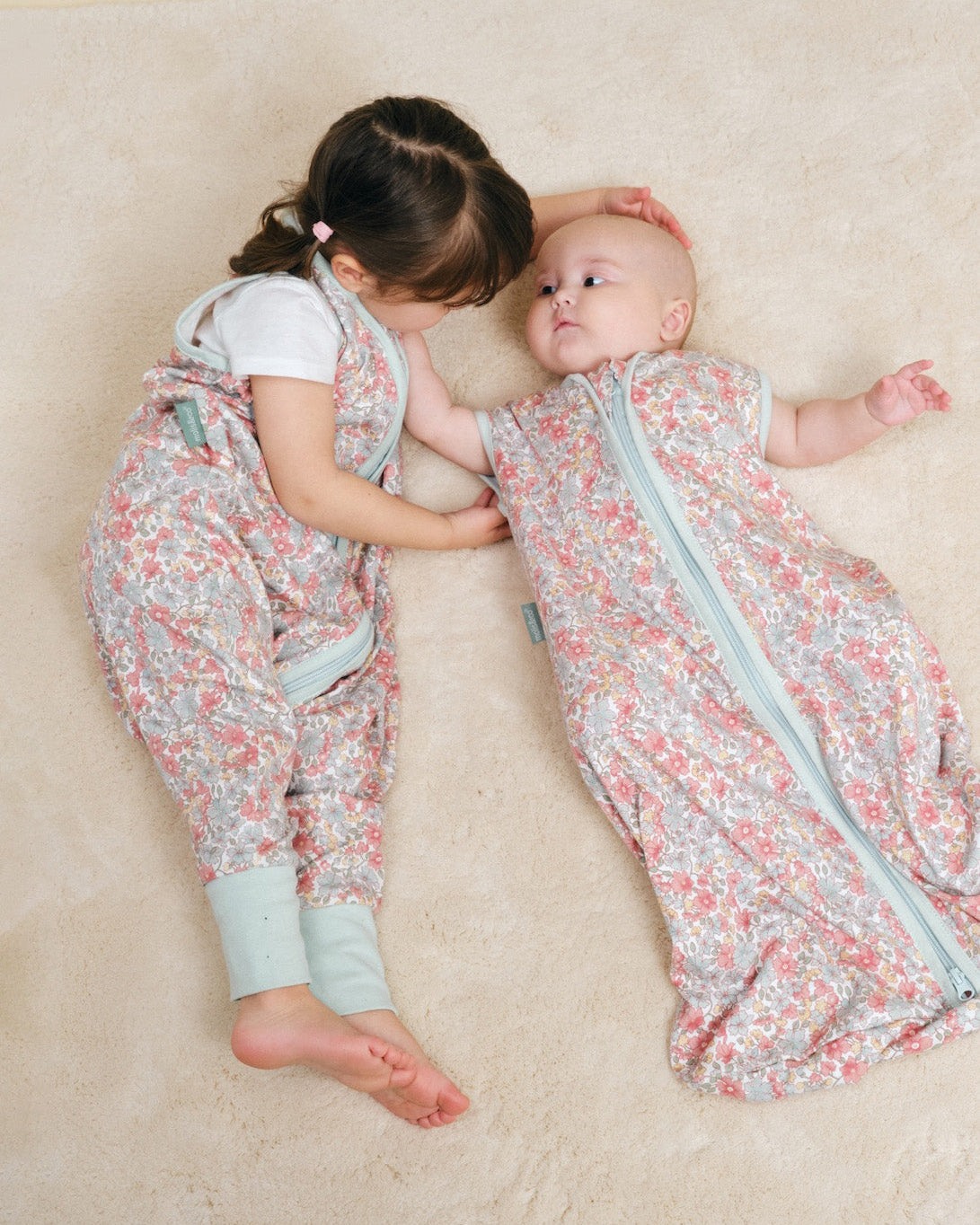 Dois bebés com roupa floral de saco de dormir e calças deitadas num tapete claro