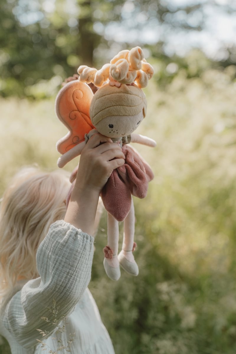 Boneca de pelúcia fada em tons de bege, rosa e laranja, segurada por criança com roupa branca ao ar livre