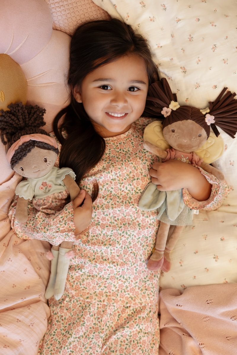 Menina deitada na cama segurando duas bonecas de pano com roupa colorida e flores.
