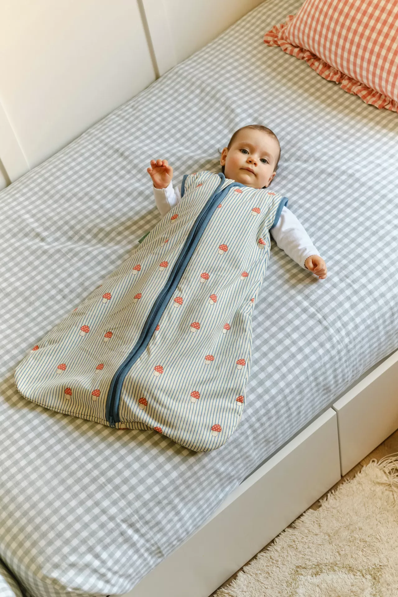 Bebé deitado com saco de dormir padrão cogumelos e riscas em cama com lençol cinzento às riscas