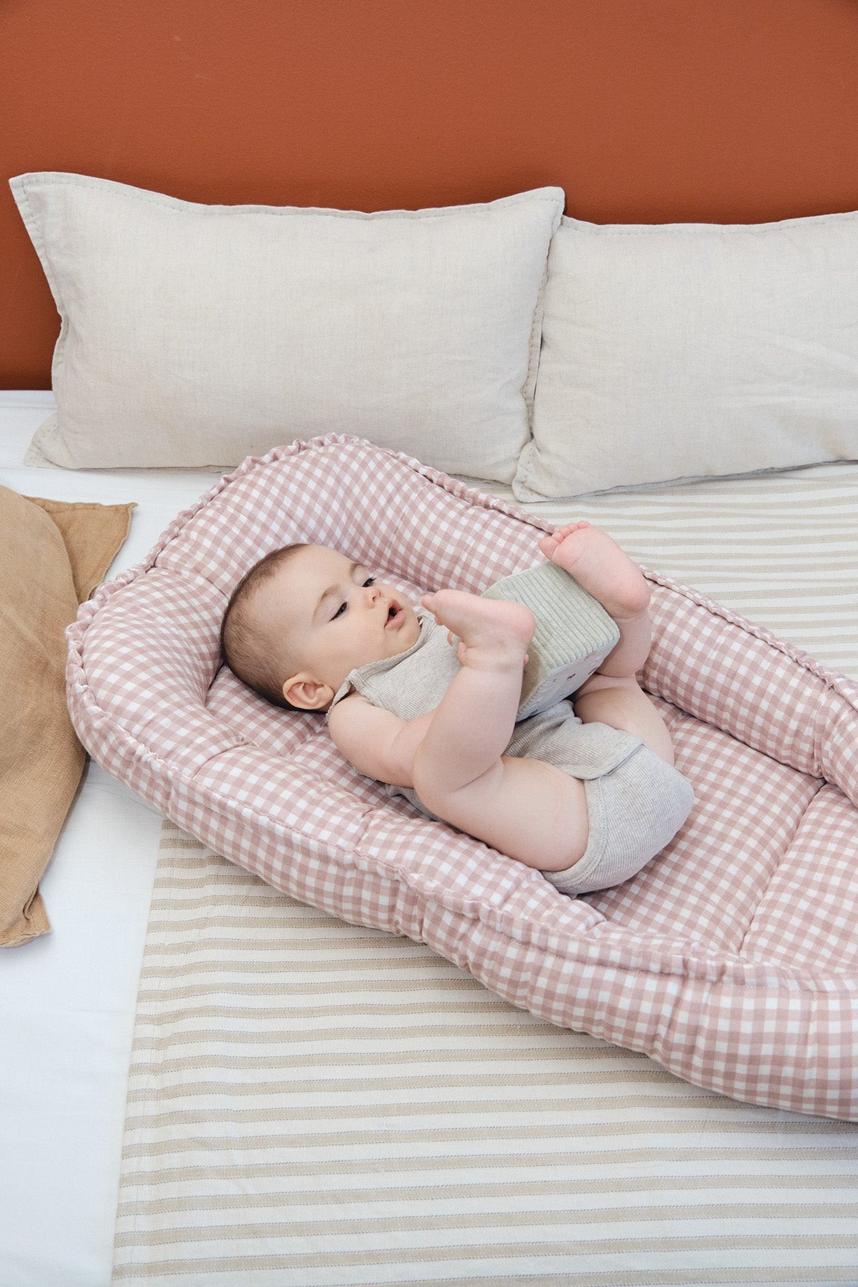 Berço portátil xadrez rosa e branco com bebé e almofadas brancas em cama.